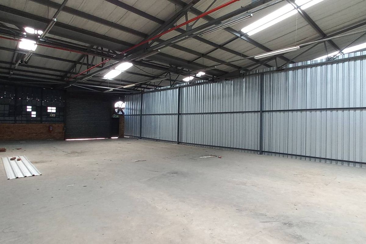 Spacious industrial warehouse unit featuring high ceilings with exposed steel trusses, corrugated metal walls, concrete flooring, and a large roller shutter door.