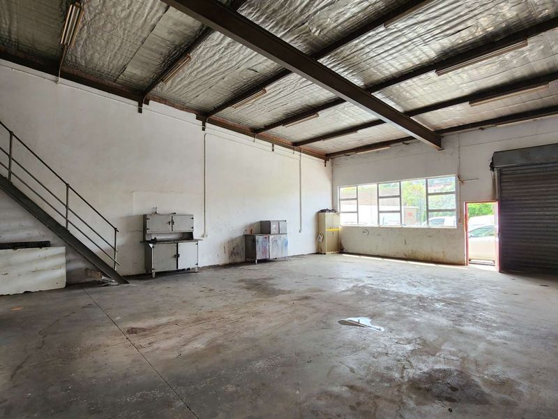 Spacious industrial warehouse or garage featuring high ceilings with exposed steel beams, concrete flooring, a metal staircase, and a large roller shutter door.
