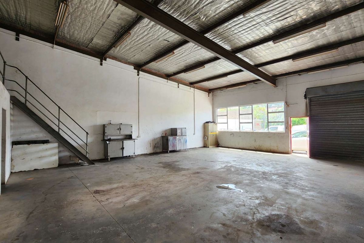 Spacious industrial warehouse or garage featuring high ceilings with exposed steel beams, concrete flooring, a metal staircase, and a large roller shutter door.
