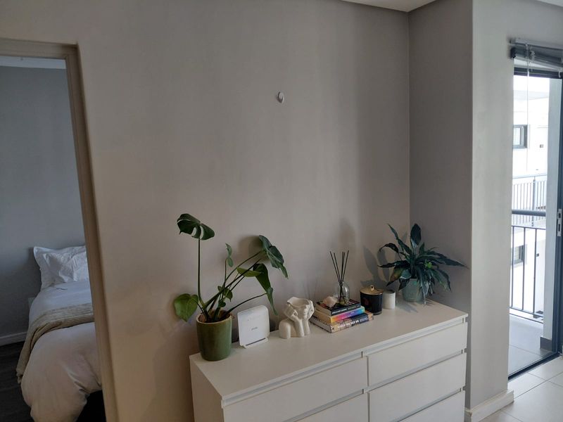 A bright living area featuring neutral walls, a sliding glass door leading to a balcony, and a view into an adjacent bedroom.