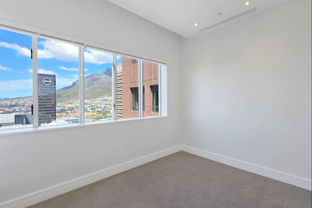 A bright, empty bedroom featuring neutral carpeting, clean white walls, and a large window offering expansive city and mountain views.