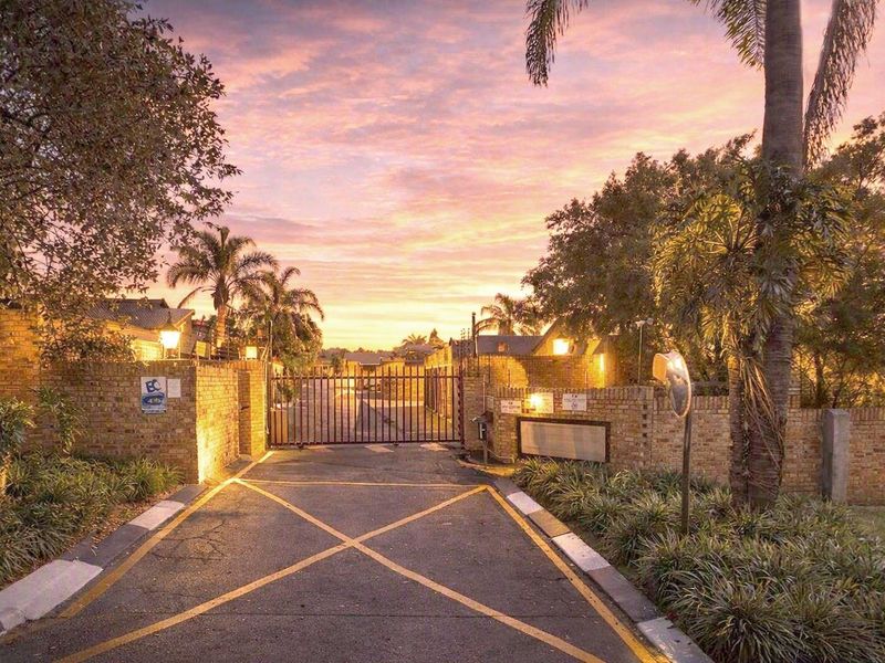 A secure gated entrance to the townhouse complex featuring brick boundary walls, asphalt driveway, and established landscaping at sunset.