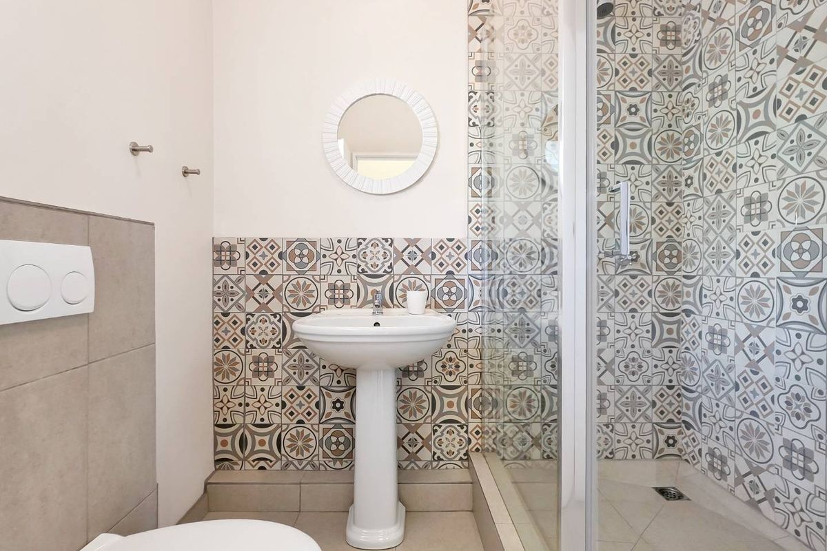 A clean, modern bathroom featuring a pedestal sink, wall-mounted toilet, and a glass-enclosed shower with decorative patterned tiling.