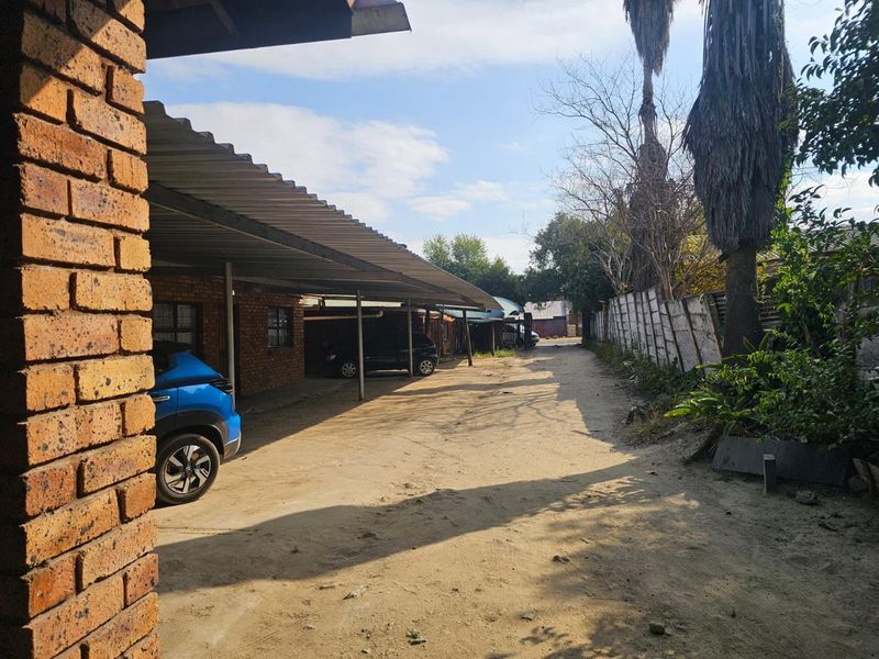 Rear exterior view showing a long carport structure, unpaved driveway, and a leaning precast concrete boundary wall.
