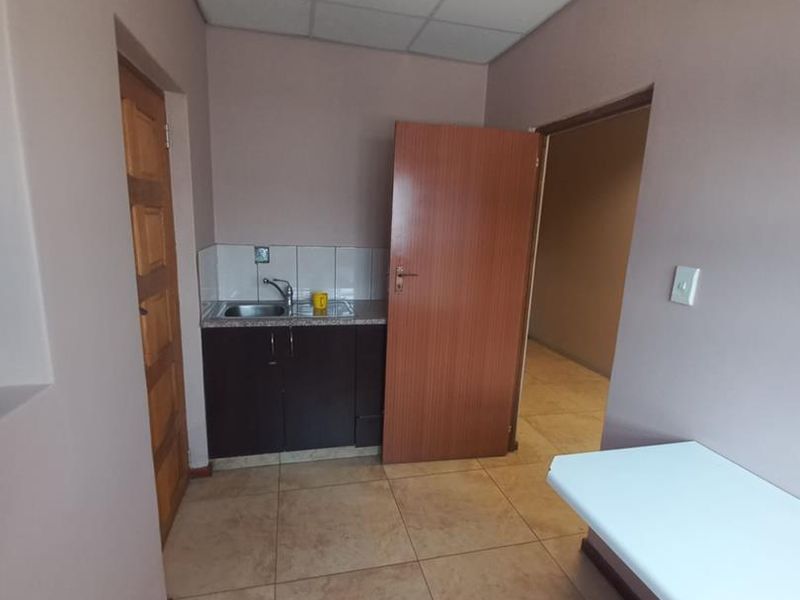 A compact kitchenette area featuring dark cabinetry, a stainless steel sink, tiled backsplash, and neutral tiled flooring within a functional office or utility space.
