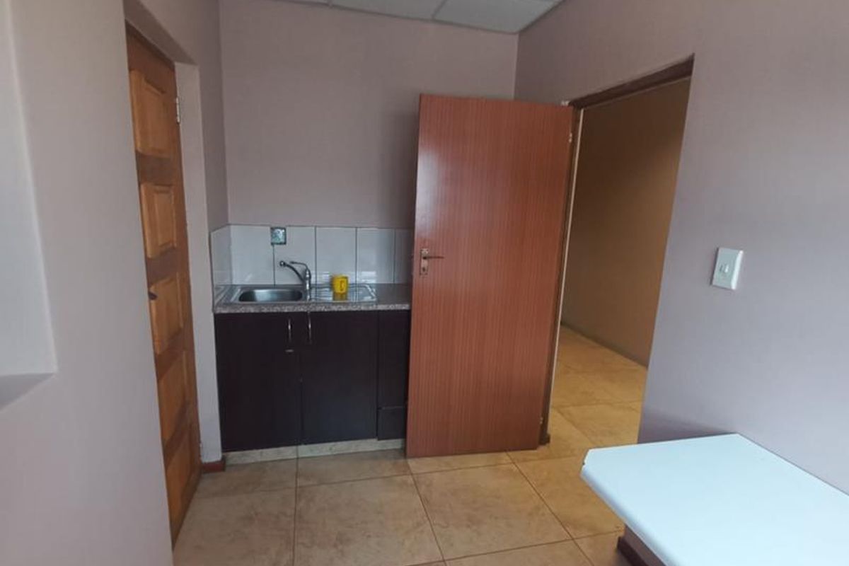 A compact kitchenette area featuring dark cabinetry, a stainless steel sink, tiled backsplash, and neutral tiled flooring within a functional office or utility space.