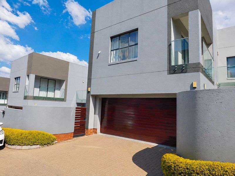 Modern double-storey cluster home featuring a dark wood garage door, paved driveway, and contemporary glass balcony balustrades.