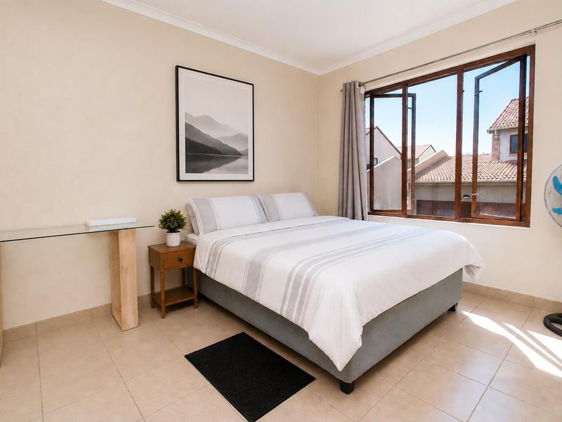 A well-lit bedroom featuring neutral tiled flooring, cove cornicing, and large casement windows providing natural light and views of the surrounding townhouse complex.
