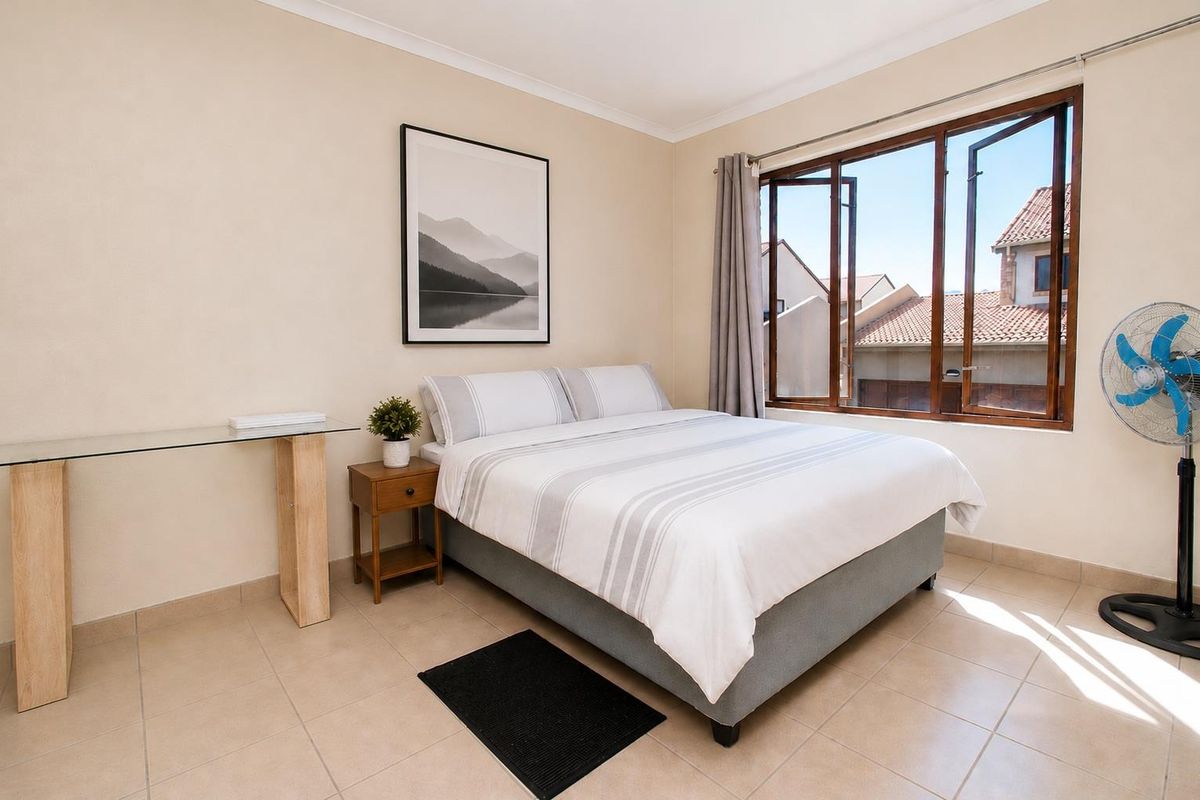 A well-lit bedroom featuring neutral tiled flooring, cove cornicing, and large casement windows providing natural light and views of the surrounding townhouse complex.