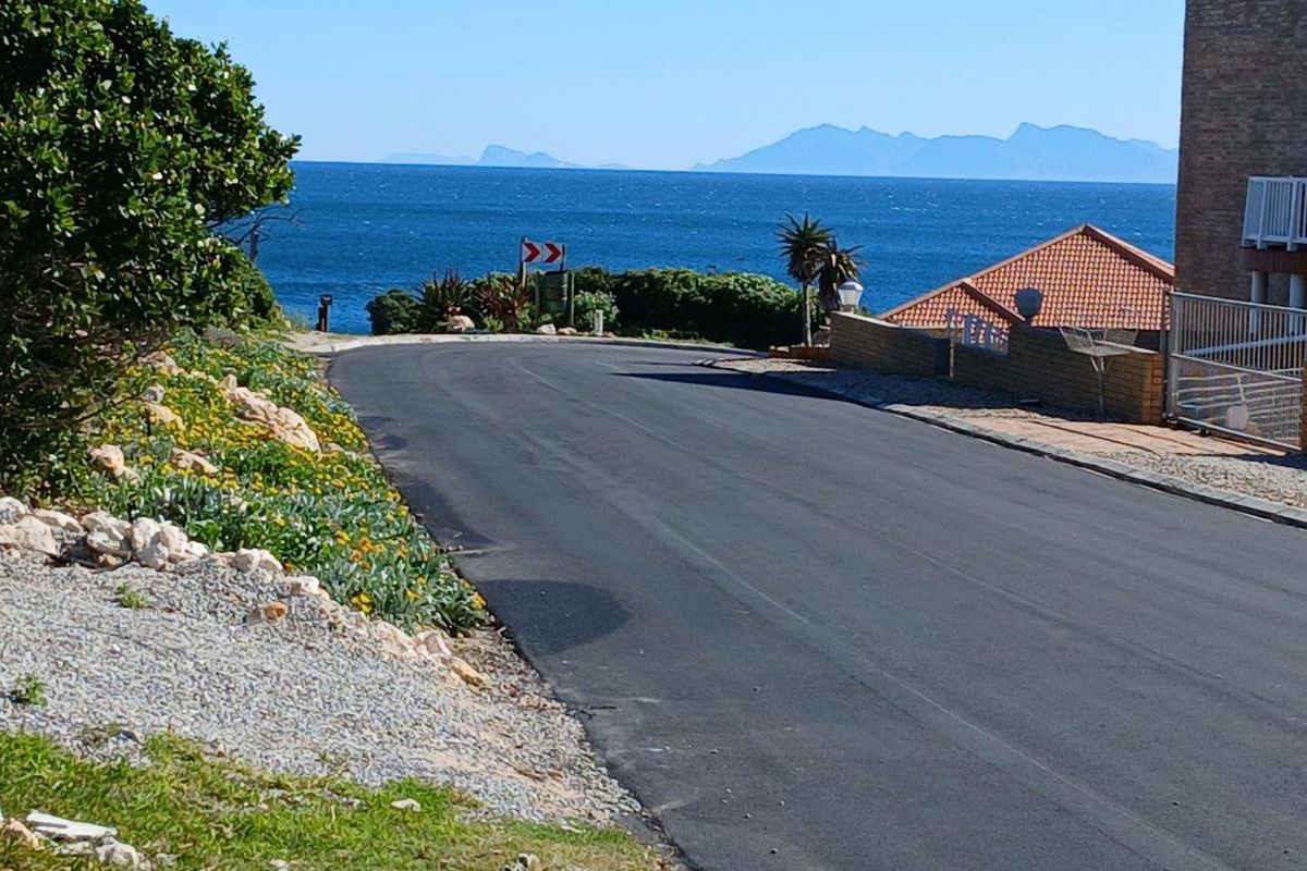 A street view of the residential area featuring a paved road, boundary wall, and scenic ocean and mountain views in the background.