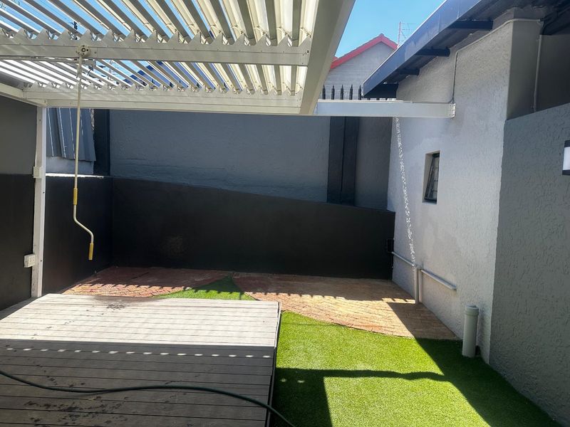A private outdoor patio area featuring a modern adjustable louvered pergola, timber decking, and a combination of paved and artificial grass surfaces.
