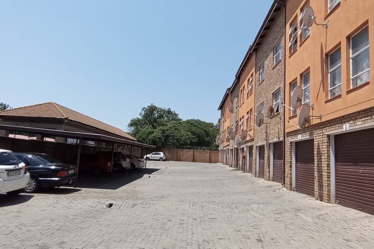 Exterior view of an apartment complex featuring a paved parking area, covered carports, and multiple ground-level roller shutter garage doors.