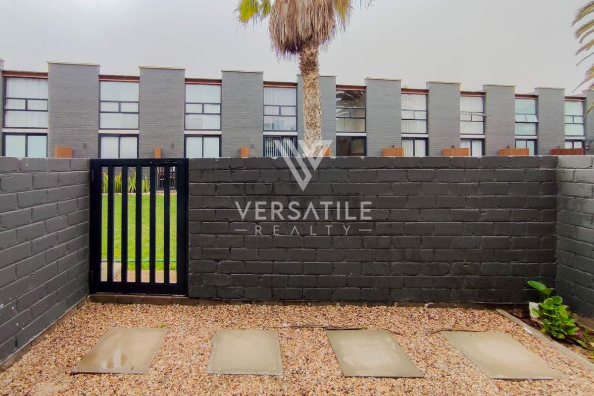 A private rear courtyard featuring a dark painted brick boundary wall, metal pedestrian gate, gravel ground cover, and concrete stepping stones.