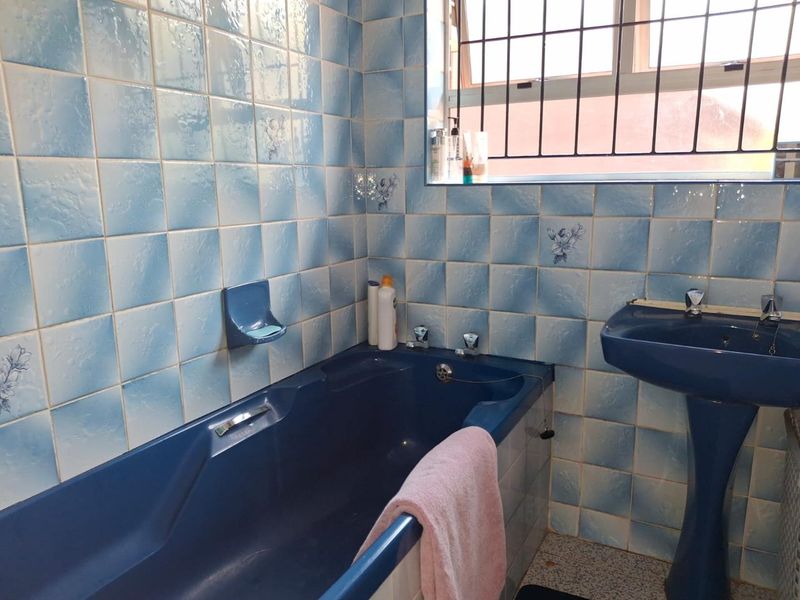 A dated bathroom featuring blue sanitaryware, matching blue patterned wall tiles, a pedestal sink, and a built-in bathtub with security bars on the window.