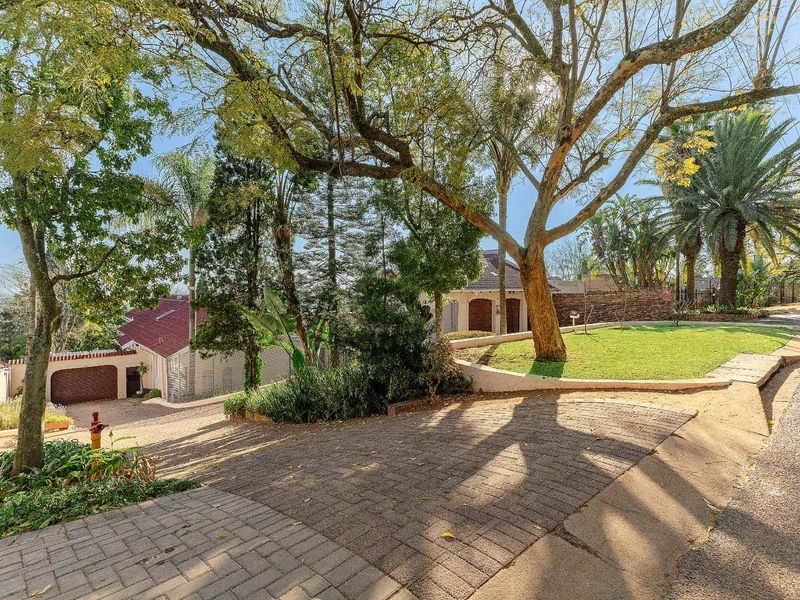 A view of the property entrance featuring a paved driveway, established trees, and a landscaped garden area.