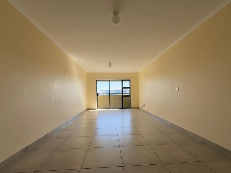 A spacious, empty living area featuring neutral tiled flooring, plain walls, and a large sliding glass door providing natural light and access to the balcony.