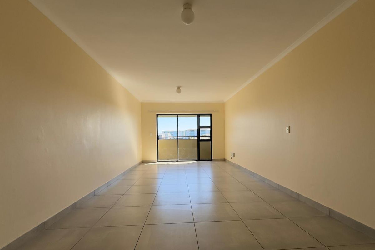 A spacious, empty living area featuring neutral tiled flooring, plain walls, and a large sliding glass door providing natural light and access to the balcony.