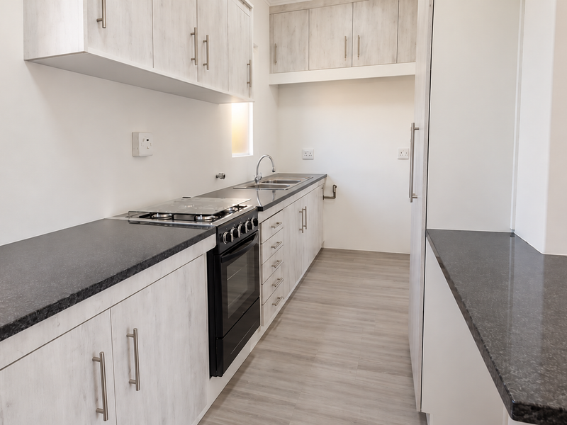 A clean, modern kitchen featuring light wood-grain cabinetry, dark laminate countertops, a built-in oven with gas hob, and durable laminate flooring.
