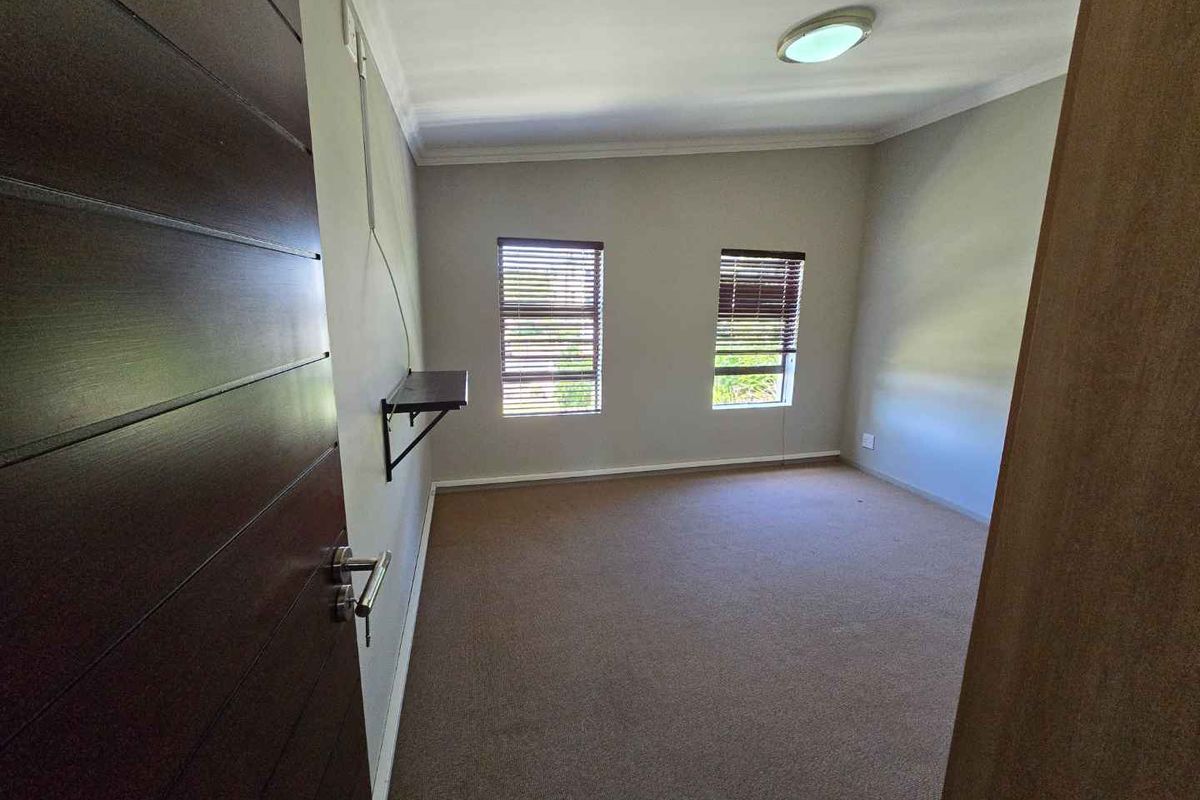 A neutral-toned bedroom featuring carpeted flooring, dual windows with fitted blinds, and a wall-mounted shelf.