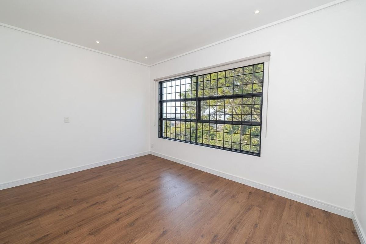 A bright, empty bedroom featuring modern wood-look laminate flooring, recessed ceiling lighting, and a large window with black security bars.