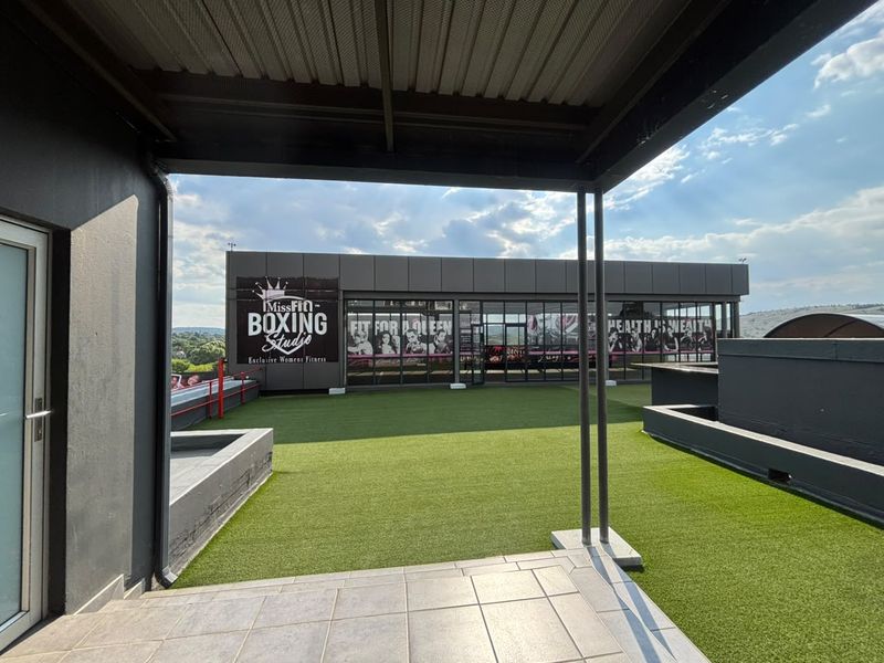 A spacious rooftop patio featuring artificial turf, tiled flooring, and a covered area with modern glass facade elements.