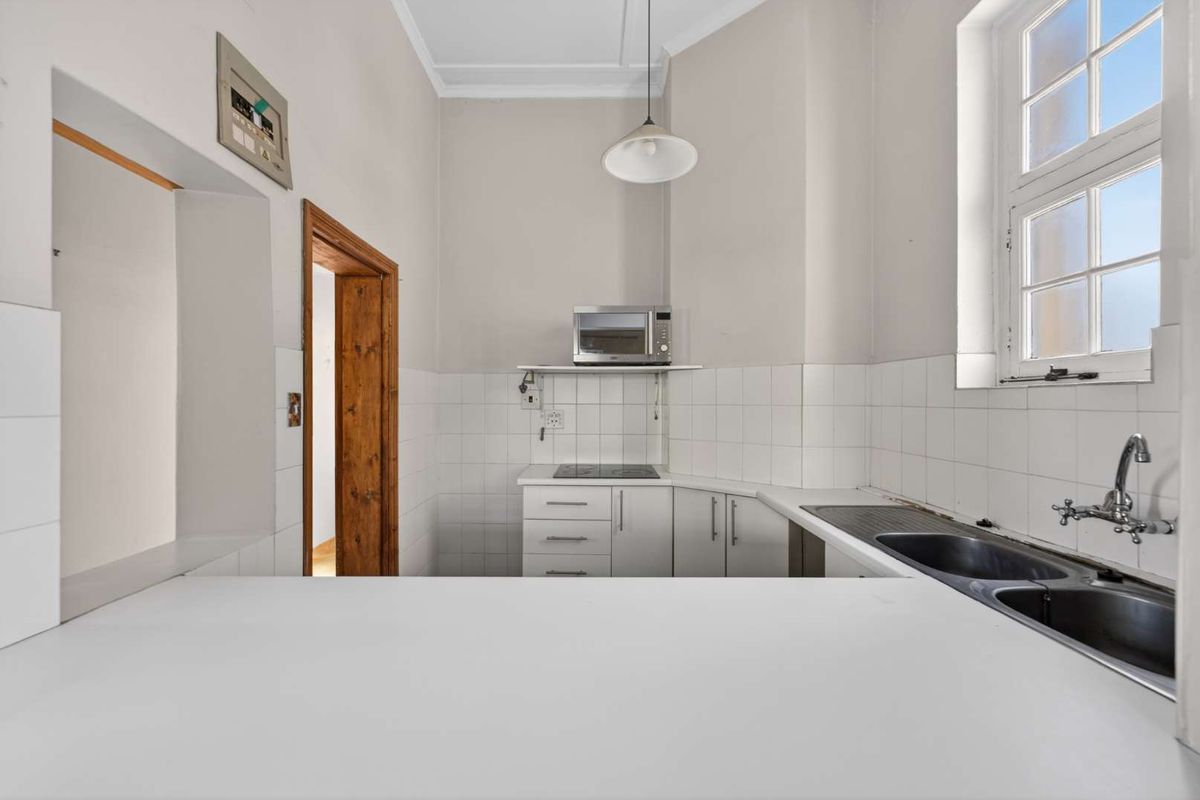 A compact, functional kitchen featuring white cabinetry, a stainless steel sink, electric hob, and tiled splashback, offering a clean canvas for potential modern updates.