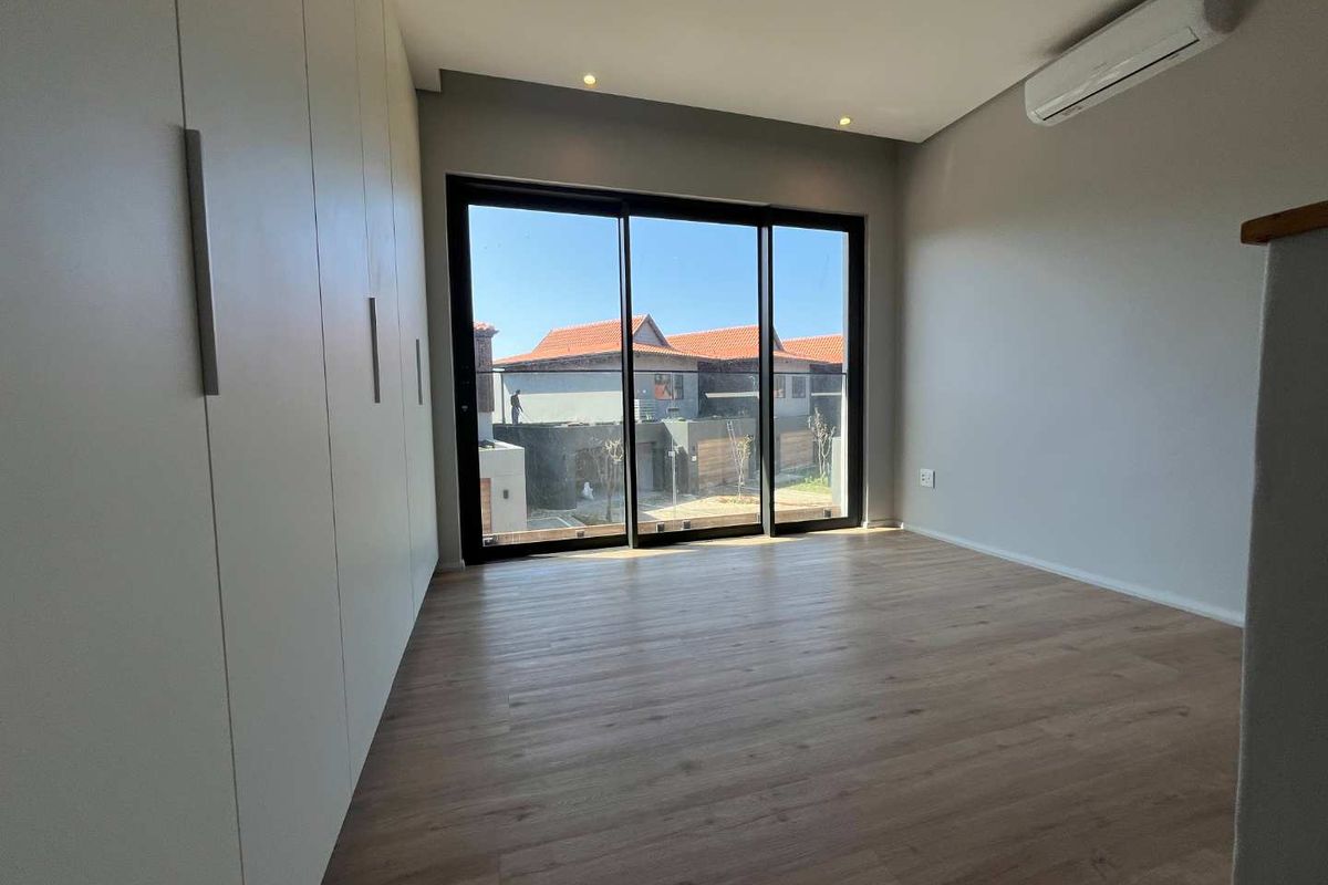 A bright, modern bedroom featuring light wood-look laminate flooring, floor-to-ceiling built-in cupboards, a split-system air conditioner, and large sliding doors leading to a balcony.