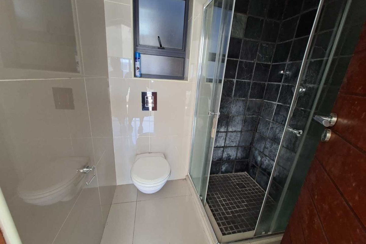 A compact bathroom featuring a glass-enclosed shower with dark textured tiling, a wall-mounted toilet, and neutral floor-to-ceiling wall tiles.
