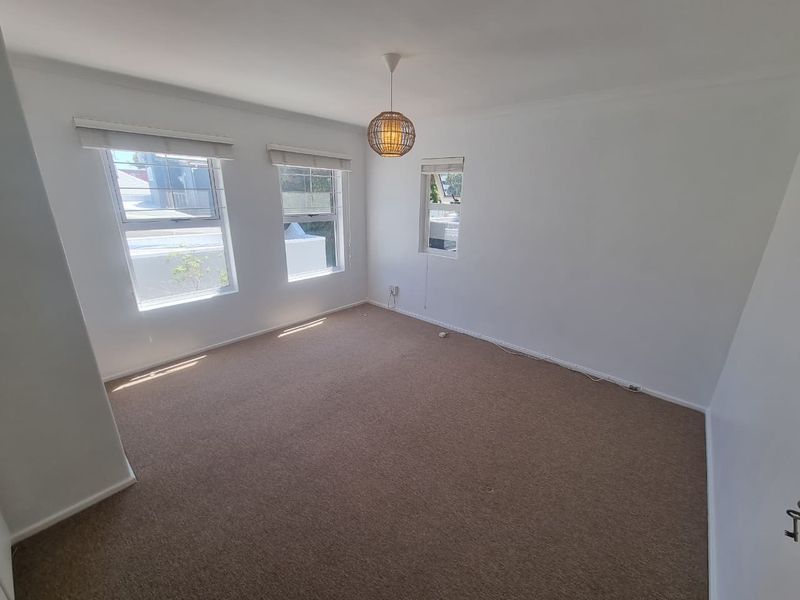 A bright, empty bedroom featuring neutral carpet flooring, multiple windows with roller blinds, and a central pendant light fixture.