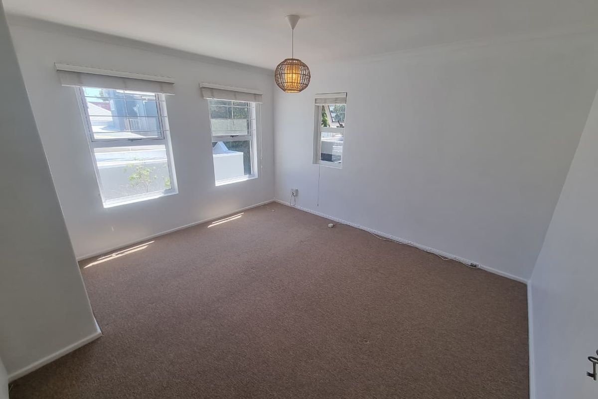 A bright, empty bedroom featuring neutral carpet flooring, multiple windows with roller blinds, and a central pendant light fixture.