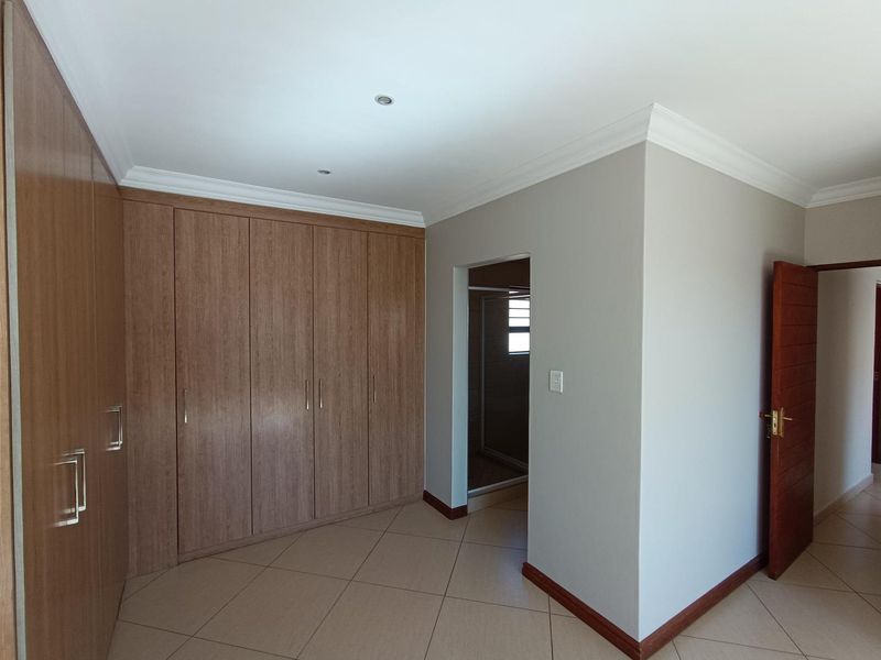 A clean, vacant bedroom featuring extensive built-in wooden cupboards, neutral tiled flooring, and coved cornicing, with an adjoining en-suite bathroom entrance.
