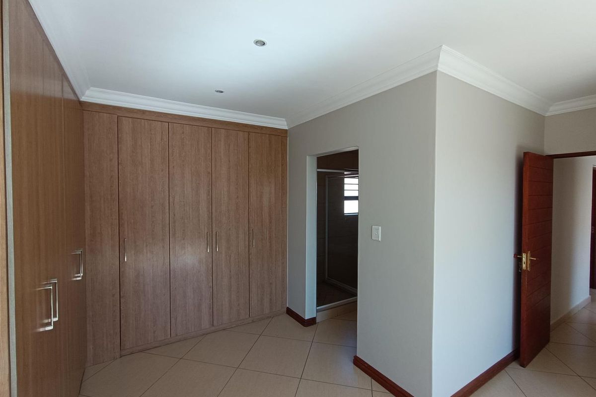 A clean, vacant bedroom featuring extensive built-in wooden cupboards, neutral tiled flooring, and coved cornicing, with an adjoining en-suite bathroom entrance.