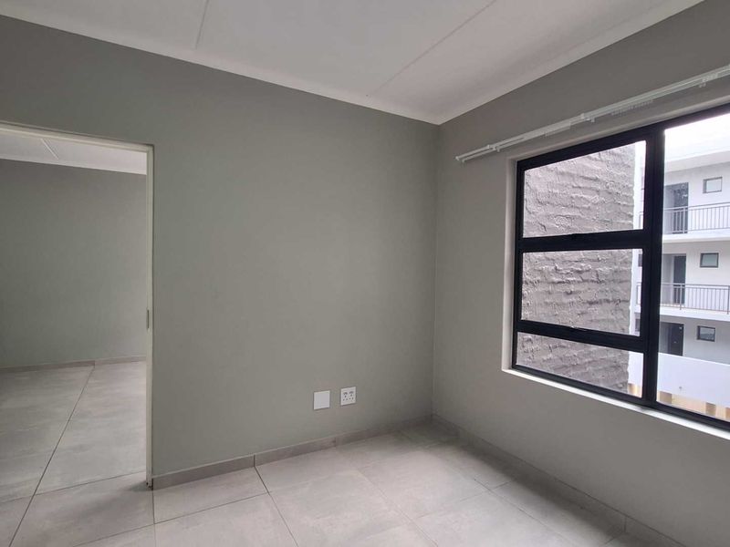 A bright, empty bedroom featuring neutral grey walls, large aluminium-framed windows, and modern tiled flooring, offering a clean and versatile living space.