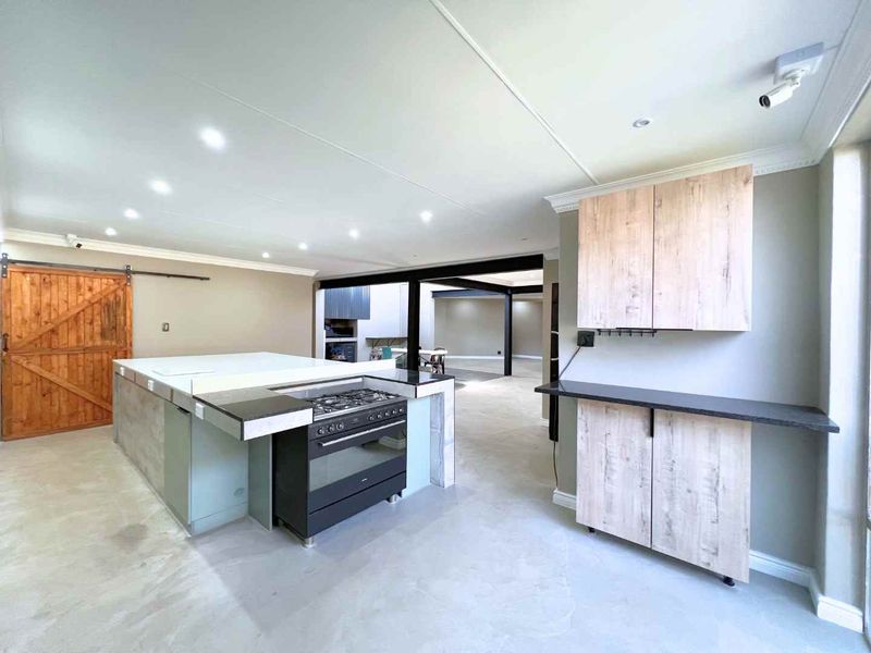 Modern open-plan kitchen featuring a large central island with a gas stove, light wood cabinetry, and industrial-style exposed steel beams.