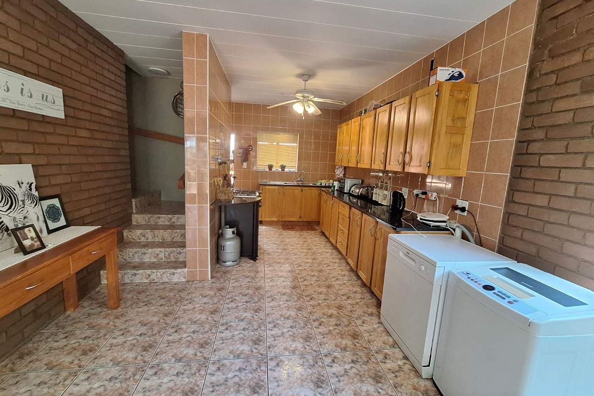 A functional kitchen featuring wooden cabinetry, dark countertops, tiled walls and flooring, with an adjacent staircase and integrated laundry appliance space.