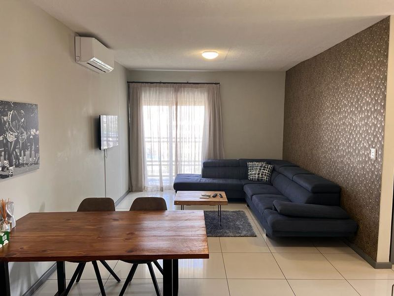 Open-plan living and dining area featuring tiled flooring, a wall-mounted air conditioning unit, and a decorative feature wall with access to a balcony.