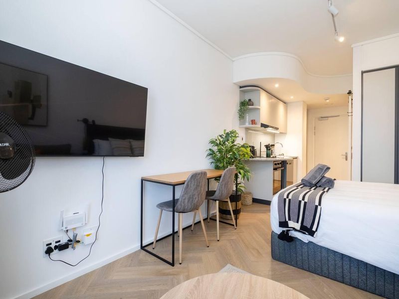 A modern open-plan studio apartment featuring herringbone flooring, a compact kitchenette, and built-in storage, offering a functional and contemporary living space.