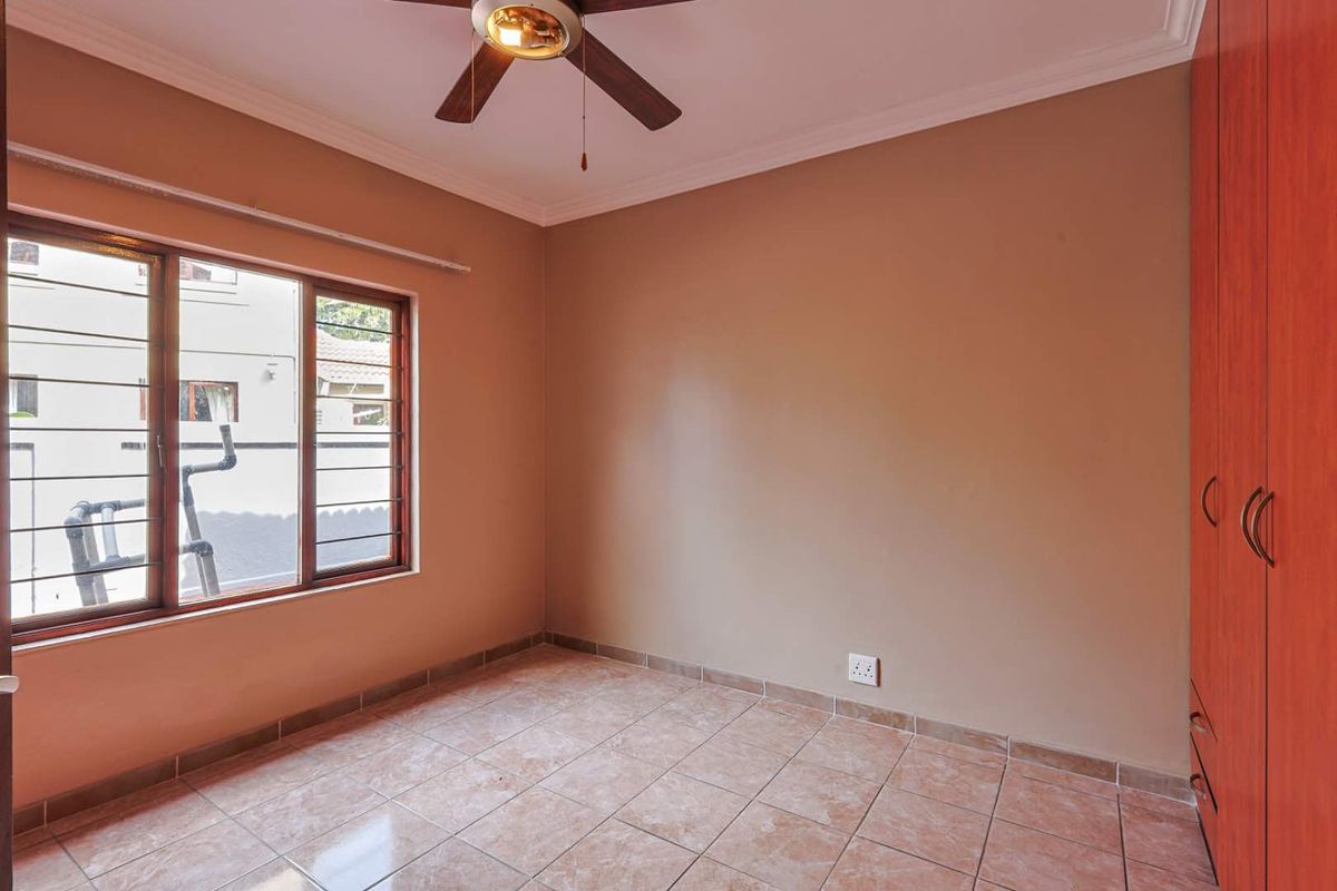 A bright, empty bedroom featuring tiled flooring, built-in wooden cupboards, a ceiling fan, and large windows providing natural light.