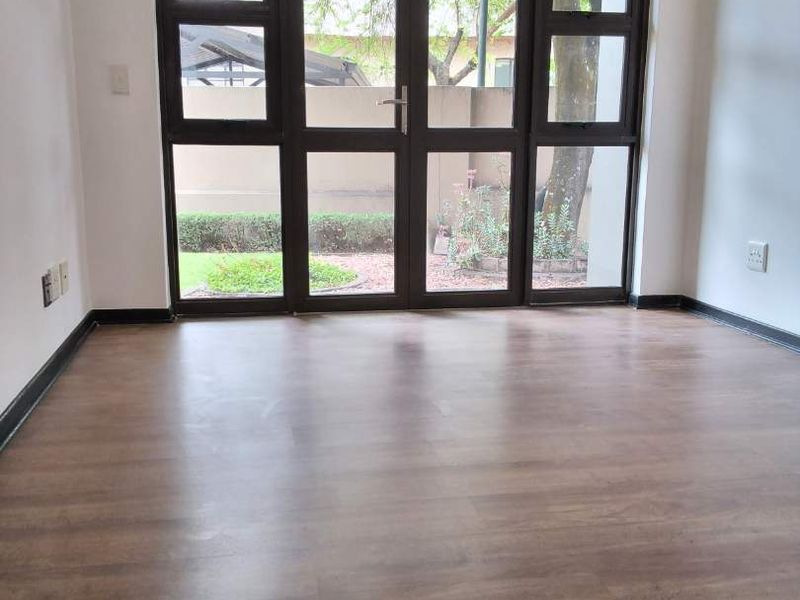A bright, empty living room featuring modern laminate flooring and large glass panel doors that provide natural light and access to the garden.