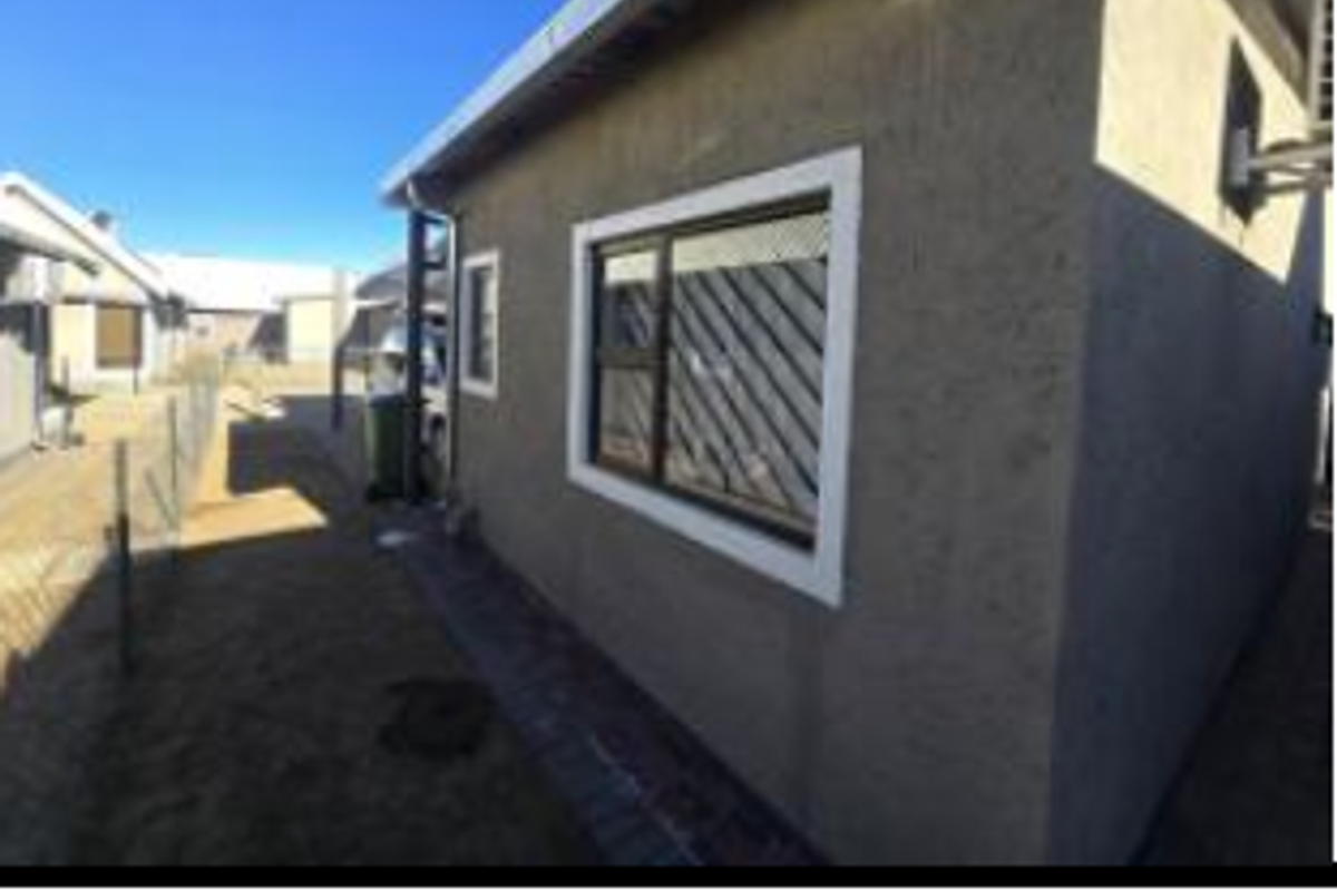 Side elevation of the house featuring textured exterior walls, aluminium window frames with security bars, and a paved walkway along the perimeter.