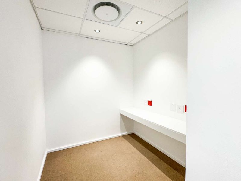 A clean, minimalist office space featuring a built-in desk, suspended ceiling with recessed lighting, and neutral carpet flooring.