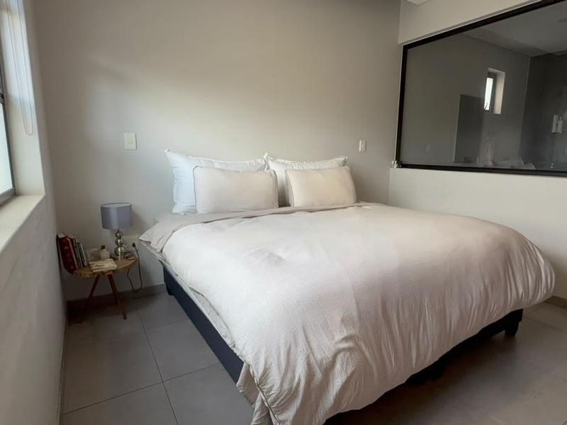 A modern bedroom featuring neutral tiled flooring, an internal glass partition wall, and large windows allowing for natural light.