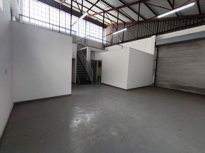 Spacious industrial warehouse unit featuring a roller shutter door, mezzanine level with steel staircase, and high ceilings with exposed roof trusses.