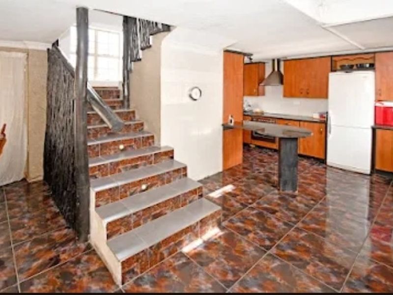Open plan kitchen and living area featuring a central tiled staircase, wooden cabinetry, and a breakfast bar with dated finishes throughout.