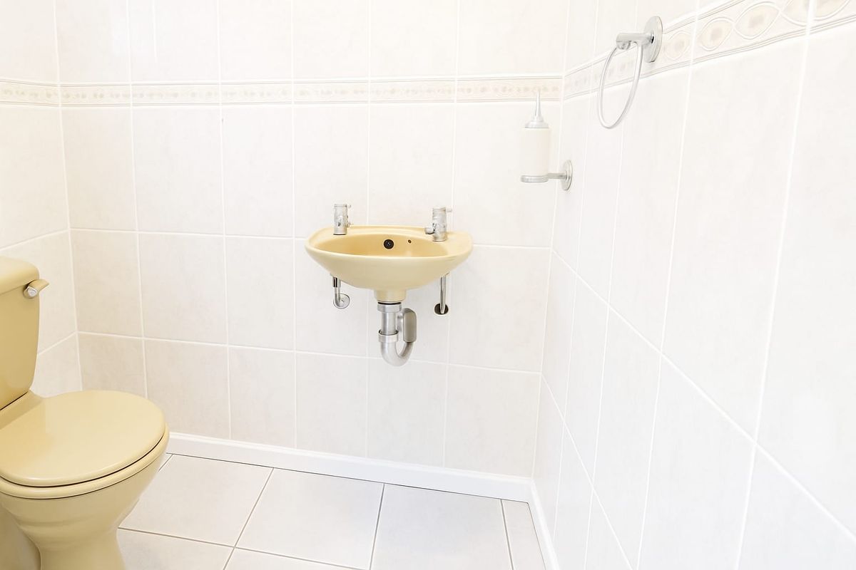 A compact bathroom featuring dated beige sanitaryware, including a wall-mounted basin and toilet, set against neutral wall and floor tiling.