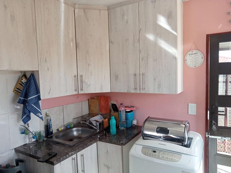A compact kitchen area featuring light wood-grain cabinetry, dark laminate countertops, a stainless steel sink, and a tiled splashback.