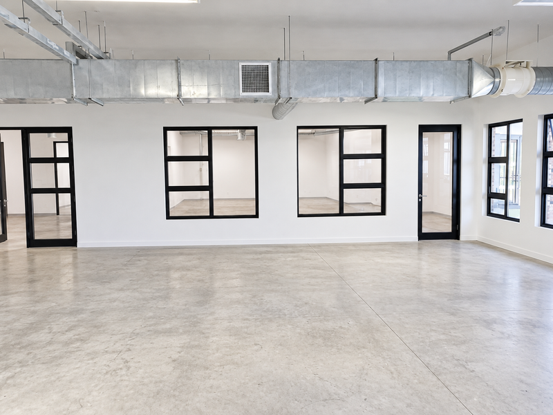 Modern open-plan industrial office space featuring polished concrete floors, exposed HVAC ducting, and sleek black-framed glass partitions providing excellent natural light and visibility.
