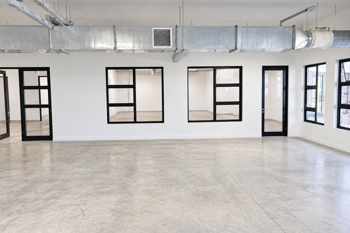 Modern open-plan industrial office space featuring polished concrete floors, exposed HVAC ducting, and sleek black-framed glass partitions providing excellent natural light and visibility.