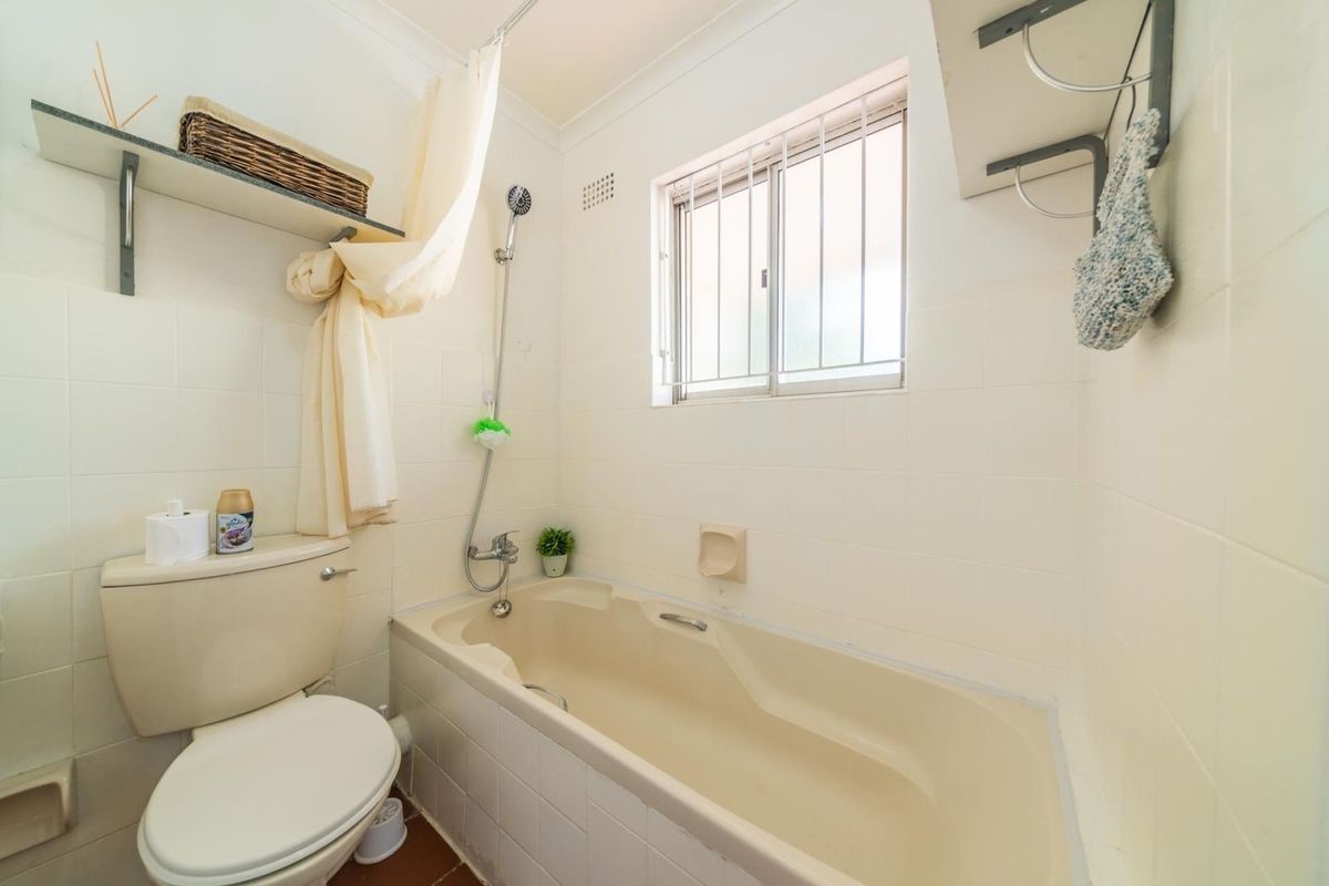 A compact bathroom featuring a bathtub with shower mixer, toilet, and wall-mounted shelving, finished with neutral wall tiles and a window with security bars.