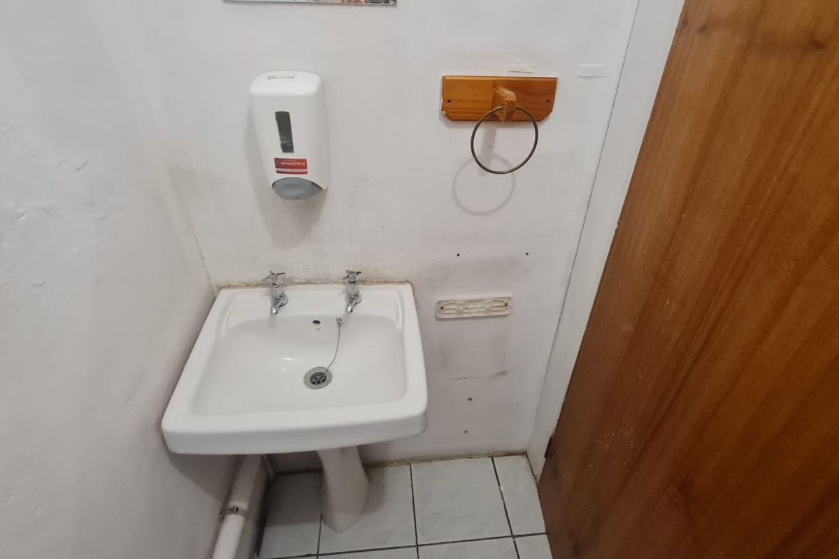 A small office washroom featuring a pedestal basin, wall-mounted soap dispenser, and towel ring, requiring cosmetic updates to the wall finishes.